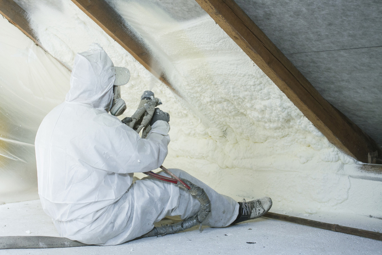 A roofer in protective gear applies spray foam insulation to the interior of a roof.