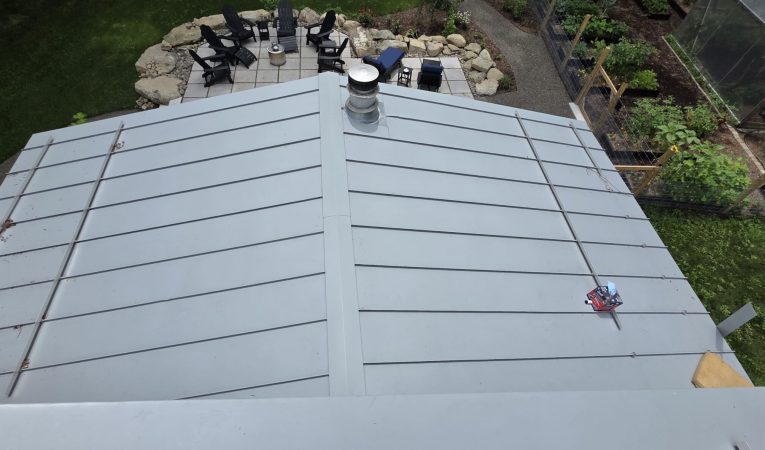 A standing seam metal roof viewed from above, with a stone patio and Adirondack chairs in view on the ground behind it, and a greenhouse and garden to the right.