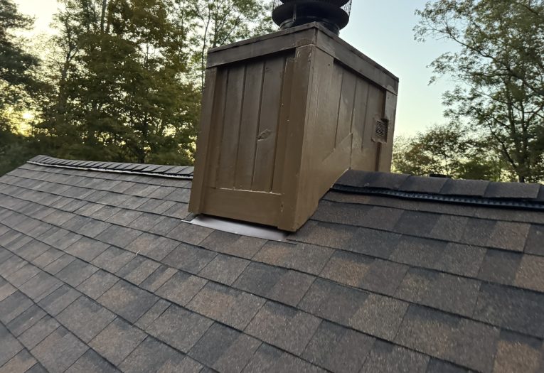 Closeup of a brown shingle roof at sunset, with a brown wood chimney cover and leafy green treetops in the background.